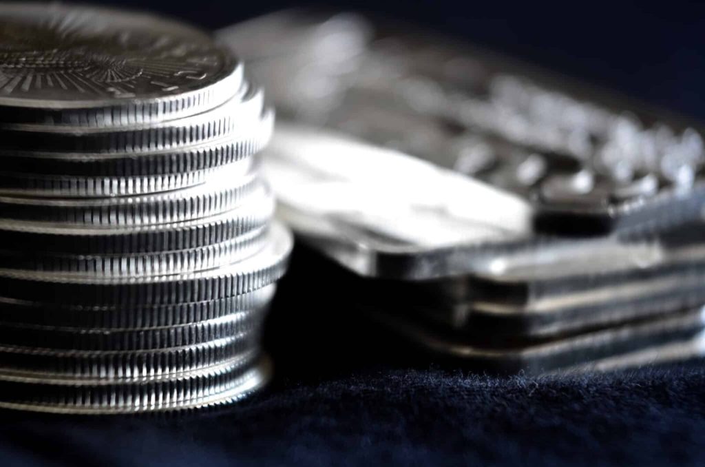 Stack of silver bullion coins beside silver bars on dark velvet surface, precious metal investment